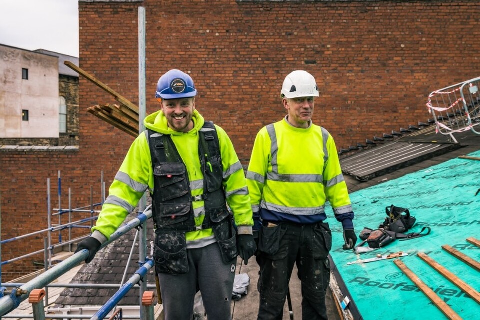 Two construction workers in high-visibility jackets and helmets stand on a scaffolding platform beside a roof under construction, with tools and building materials visible around them. One worker is smiling.